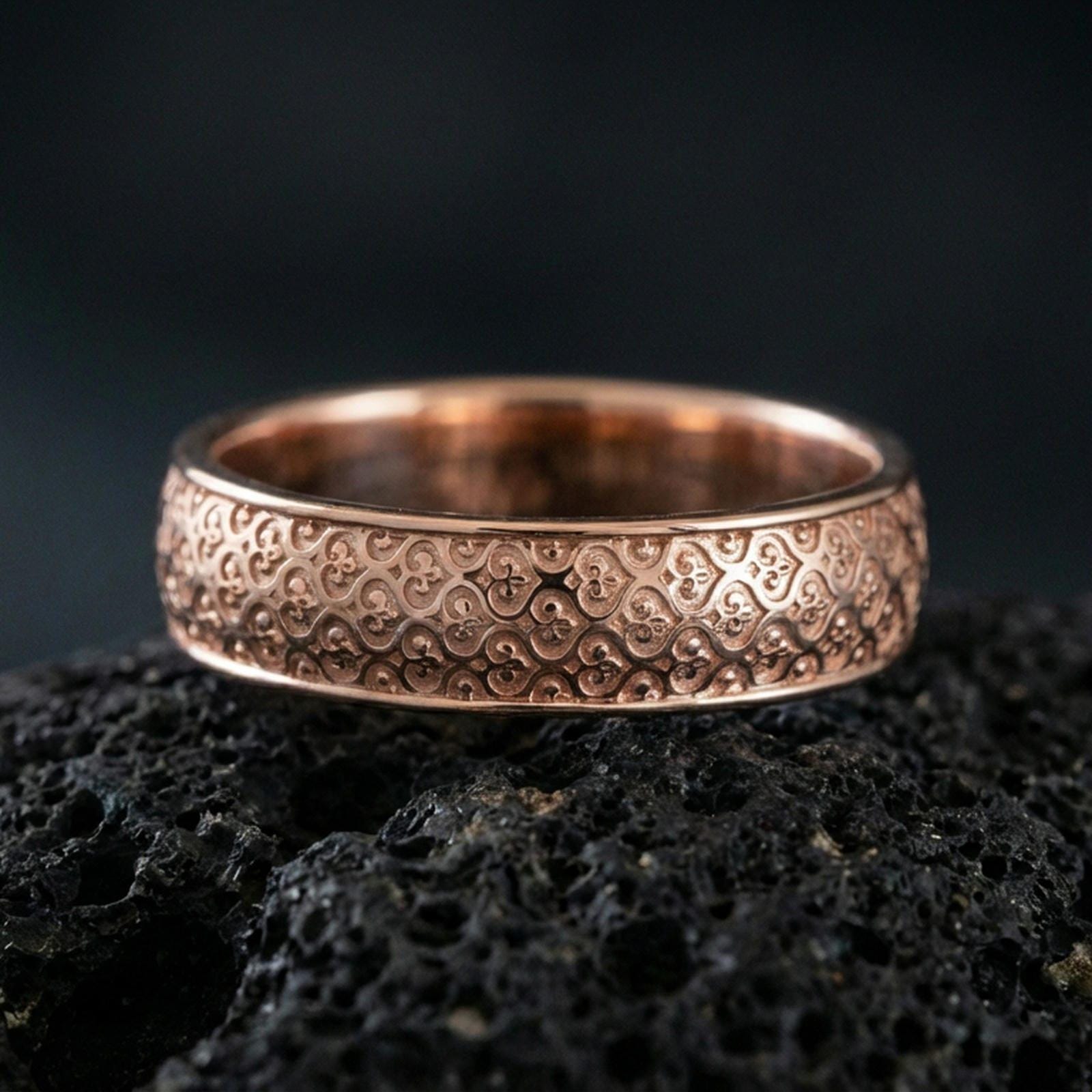 Rose gold plated fleur-de-lis band ring on volcanic stone, refined macro shot featuring soft rose highlights and sharp ornamental details.