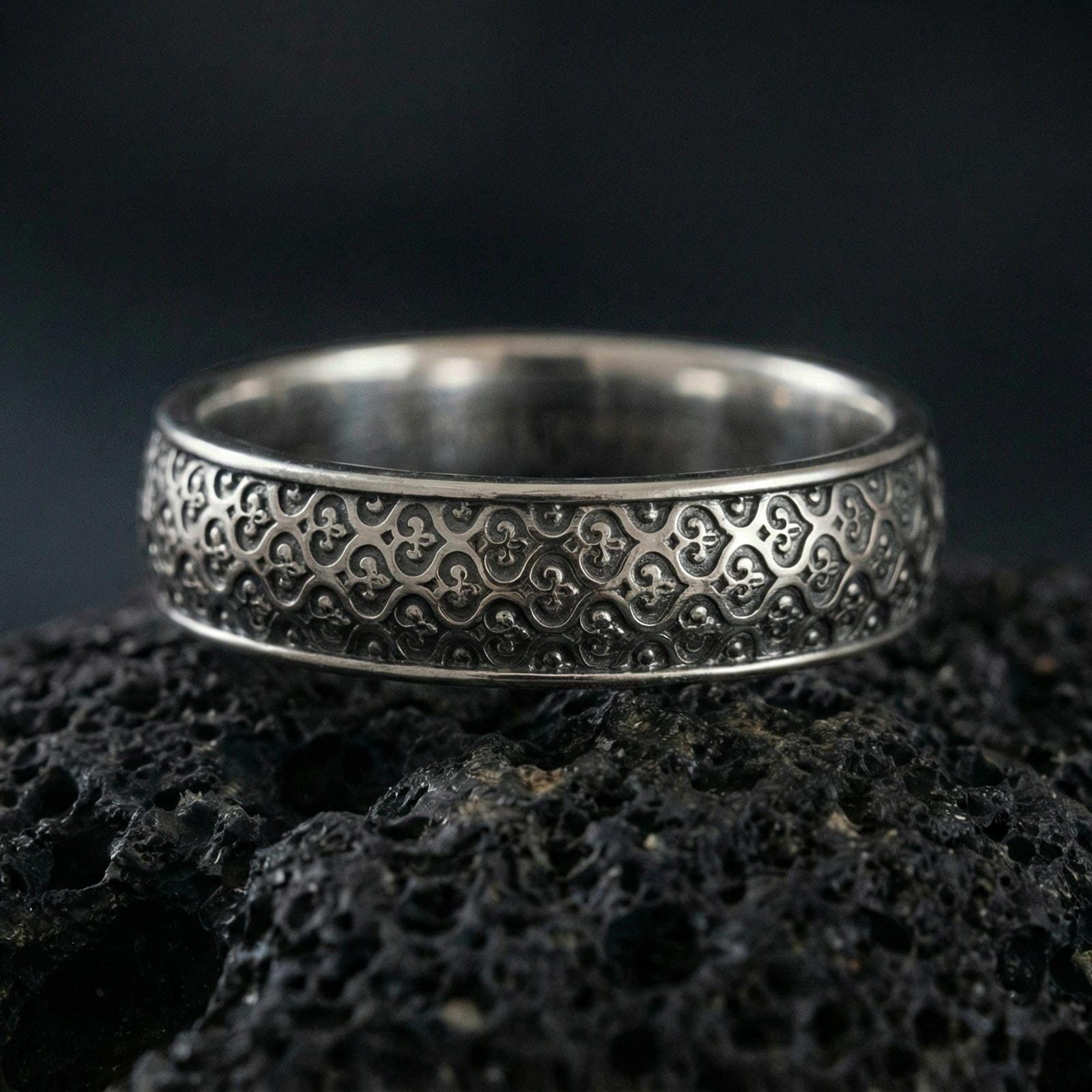 Oxidized sterling silver fleur-de-lis band ring on volcanic stone, macro close-up highlighting the repeating medieval motif and dark patina depth.