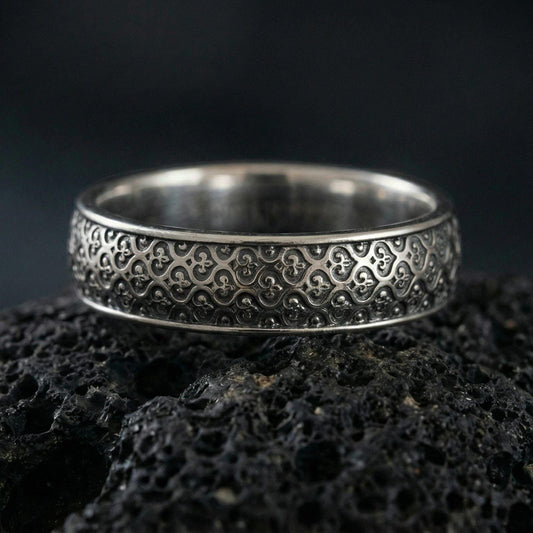 Oxidized sterling silver fleur-de-lis band ring on volcanic stone, macro close-up highlighting the repeating medieval motif and dark patina depth.