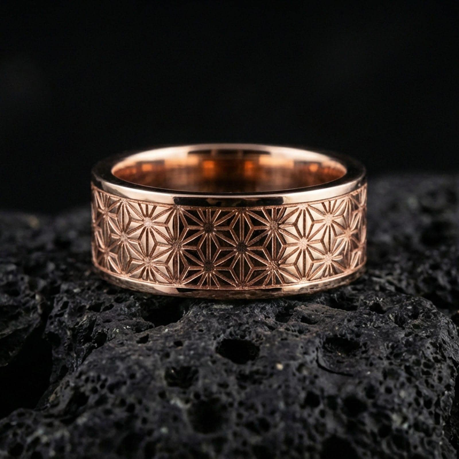 Rose gold plated geometric floral band ring on volcanic stone, refined macro shot featuring soft rose highlights and precise repeating geometric lines.