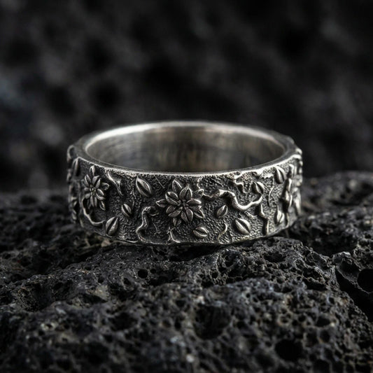 Oxidized sterling silver floral engraved band ring on volcanic stone, macro close-up highlighting raised blossoms, leaf motifs, and deep contrast texture.