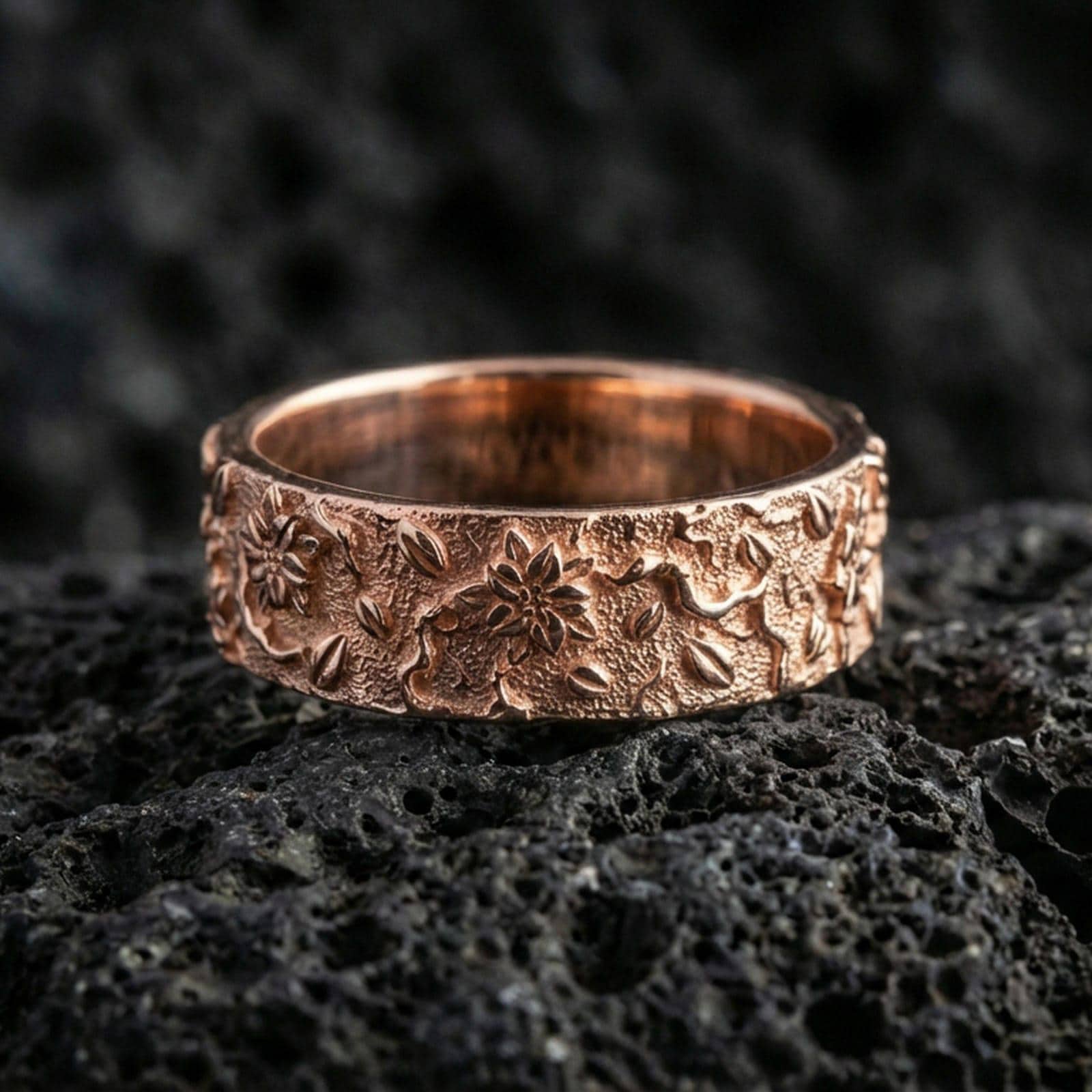 Rose gold plated floral engraved band ring on volcanic stone, elegant macro shot capturing soft rose reflections, raised petals, and refined engraved depth.