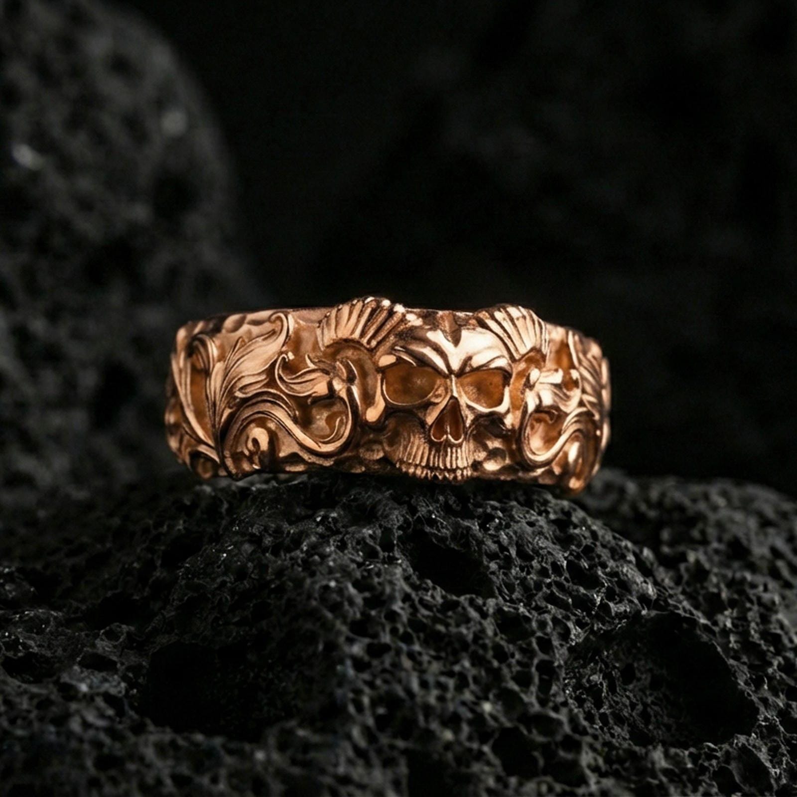 Rose gold plated skull ring on volcanic stone, refined macro close-up showcasing soft rose sheen across the skull motif and decorative scroll pattern.