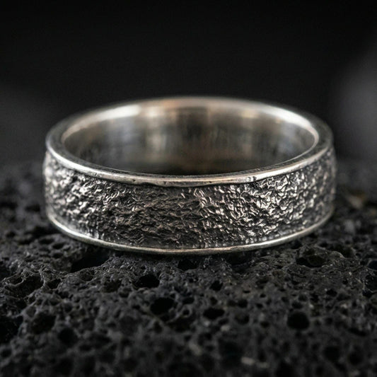 Oxidized sterling silver bark texture band ring on volcanic stone, macro close-up highlighting rugged tree-bark relief and deep shadowed contrast.