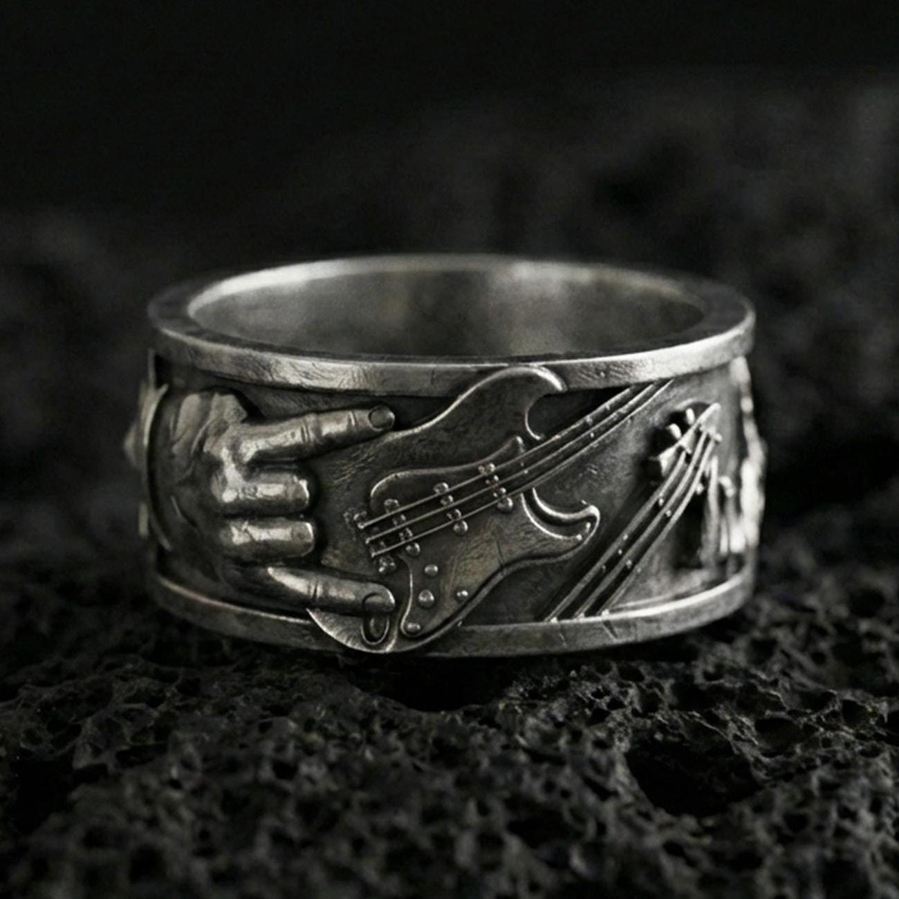 Oxidized sterling silver guitar ring on volcanic stone, macro close-up highlighting the raised electric guitar and hand detail with deep vintage patina.