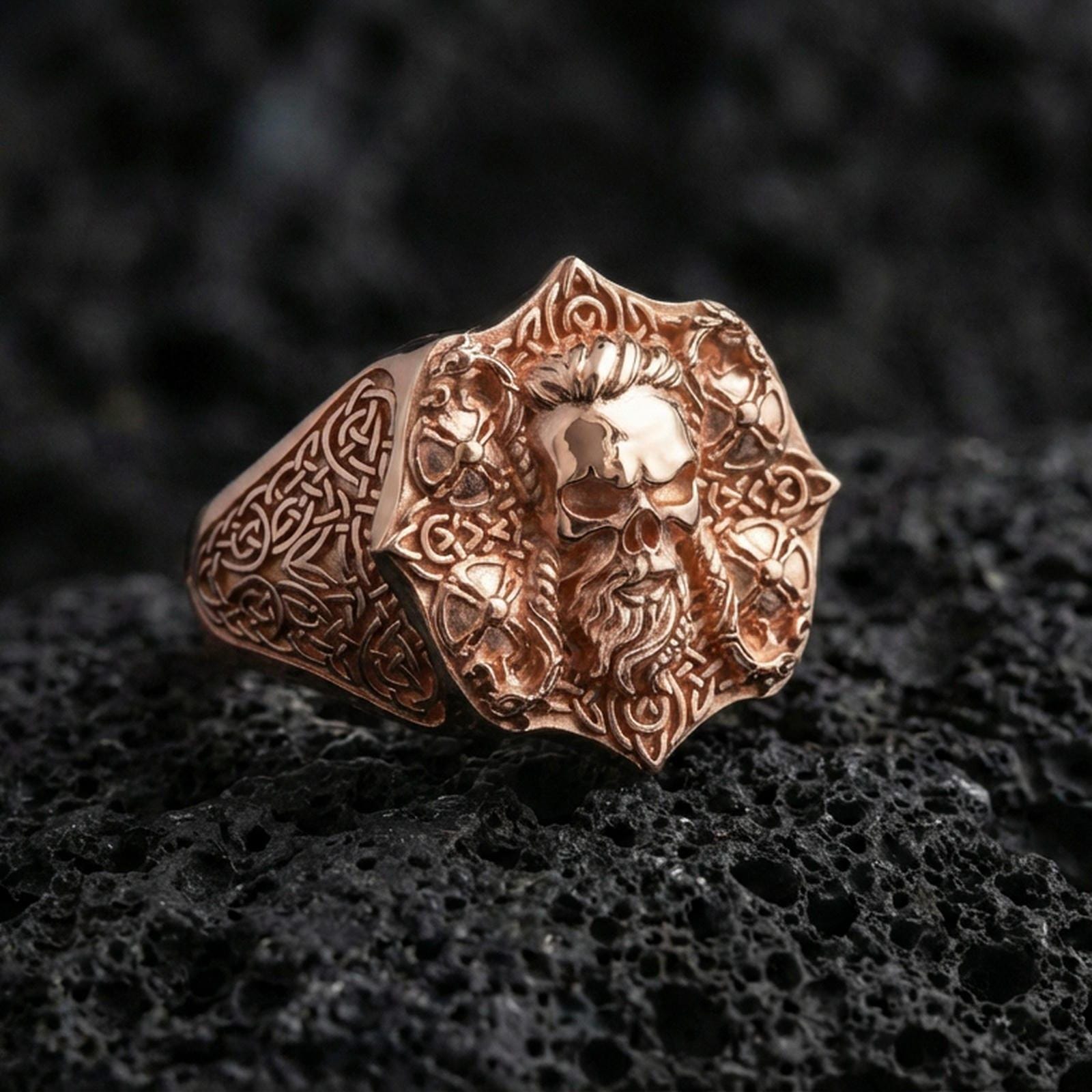 Rose gold plated skull signet ring with Celtic knot engraving on textured volcanic rock, luxury close-up capturing warm tone and sculpted skull depth