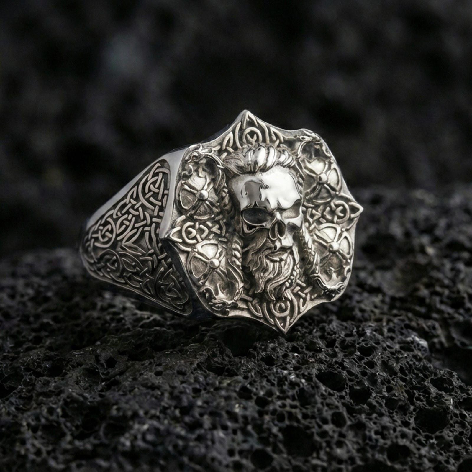Polished silver skull signet ring with intricate Celtic knotwork on porous volcanic stone, close-up highlighting sharp edges and clean reflective finish