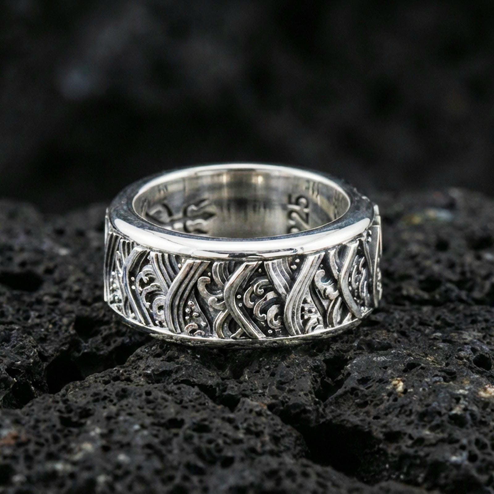 Rotating wave spinner ring on black volcanic stone, photographed in polished silver with clean highlights defining the curved wave lines and dotted texture.