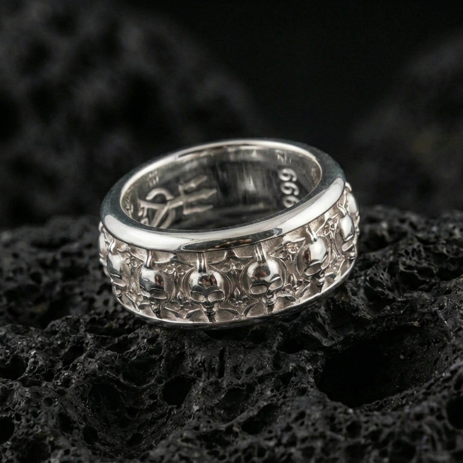Skull motif sterling silver band ring on black volcanic stone, photographed in a polished silver look with clean highlights across the raised skull pattern.