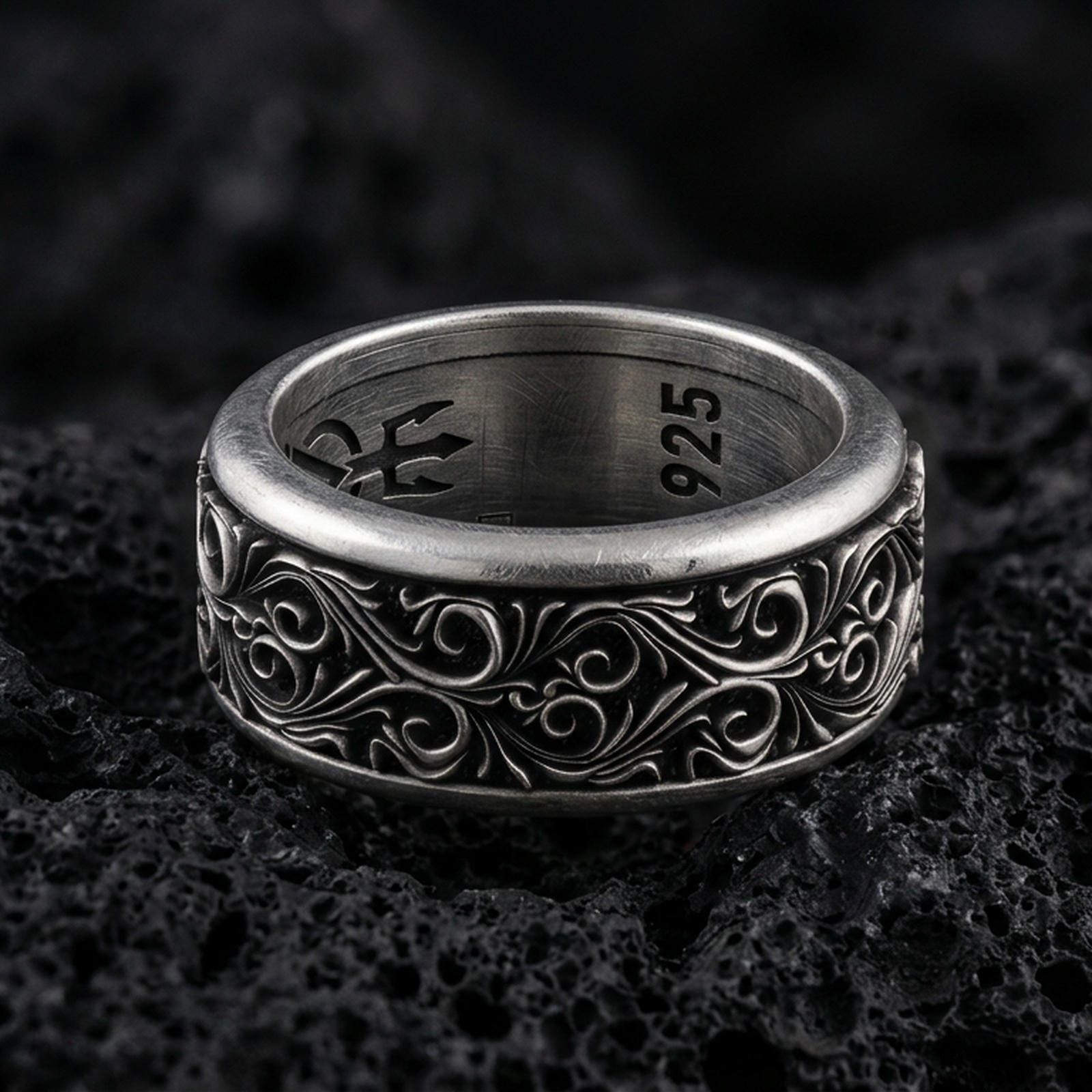 Ornate scrollwork sterling silver band ring on black volcanic stone, photographed with an oxidized finish to emphasize depth and shadow in the engraved pattern.
