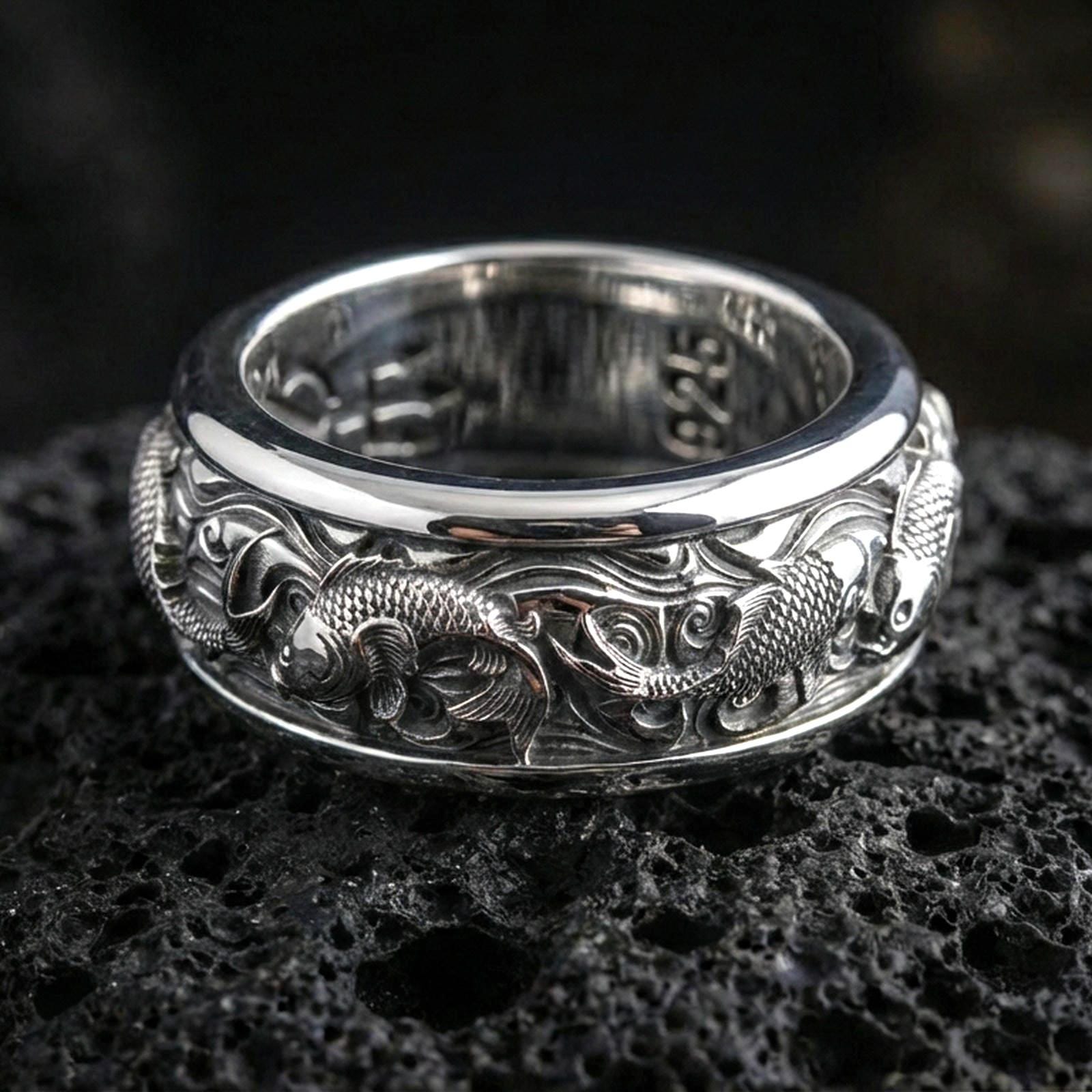 Koi fish sterling silver band ring on black volcanic stone, photographed in a polished silver look with clean highlights across the koi and wave engraving.