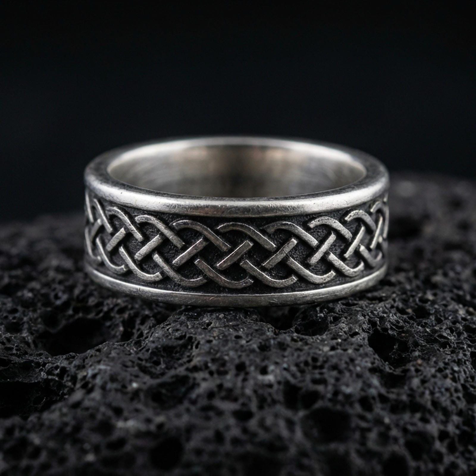 Sterling silver Celtic knot band ring on black volcanic stone, photographed with an oxidized patina that darkens the recessed background and sharpens the raised knotwork.