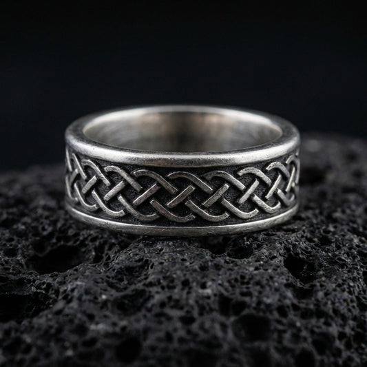 Sterling silver Celtic knot band ring on black volcanic stone, photographed with an oxidized patina that darkens the recessed background and sharpens the raised knotwork.