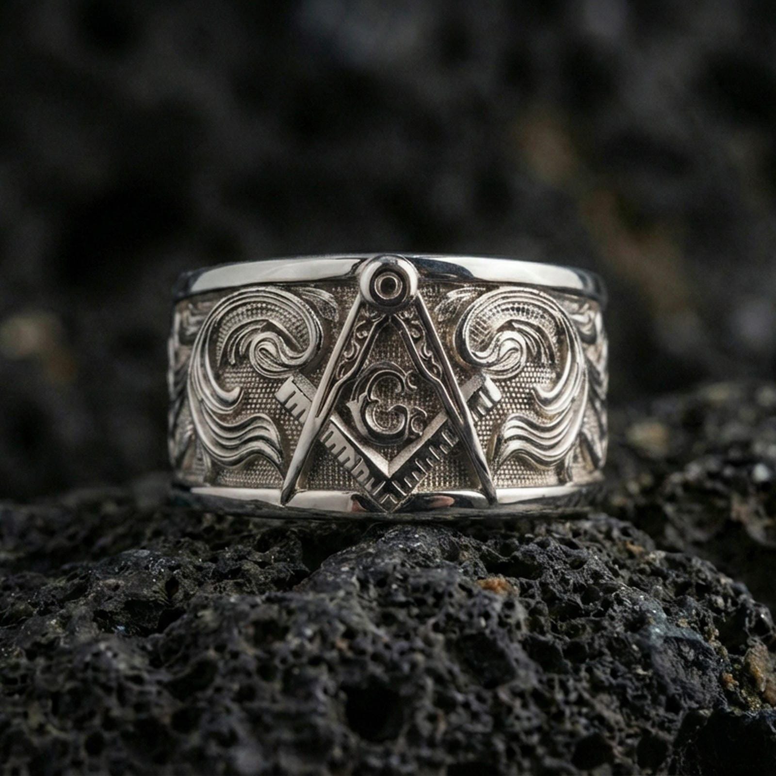 Sterling silver Masonic square and compass band ring on black volcanic stone, shown in a polished silver look with crisp reflections across the square, compass, and decorative scrollwork.