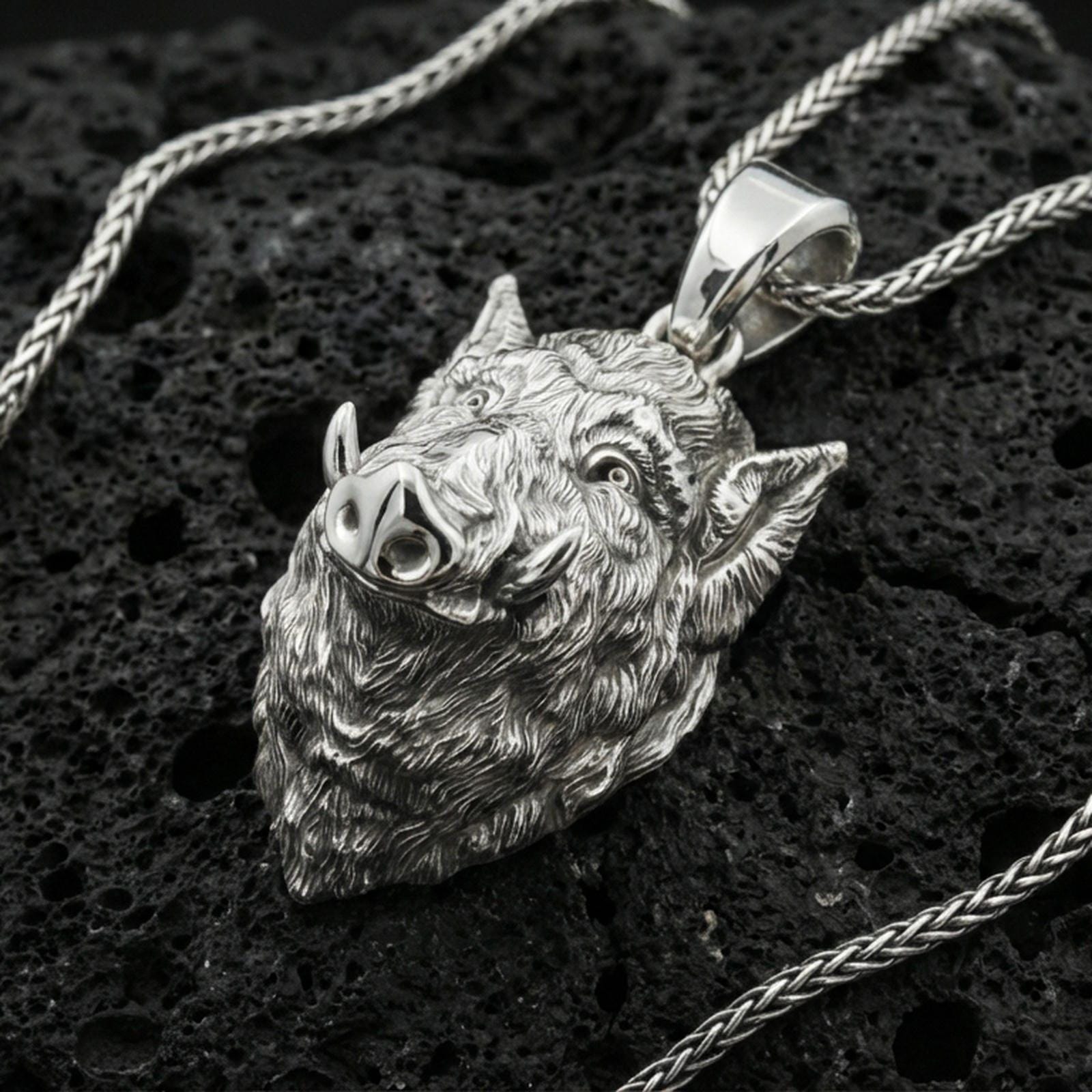 Polished silver boar head pendant on porous volcanic stone with braided chain, close-up highlighting sharp tusks and crisp sculpted details