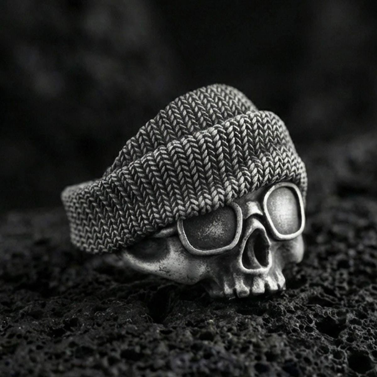 Oxidized sterling silver skull ring with beanie and goggles on black volcanic rock, macro close-up showing deep patina and streetwear texture