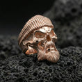 Load image into Gallery viewer, Rose gold plated bearded skull ring with ribbed beret on textured volcanic rock, luxury close-up capturing soft warm plating glow and high-relief gothic details
