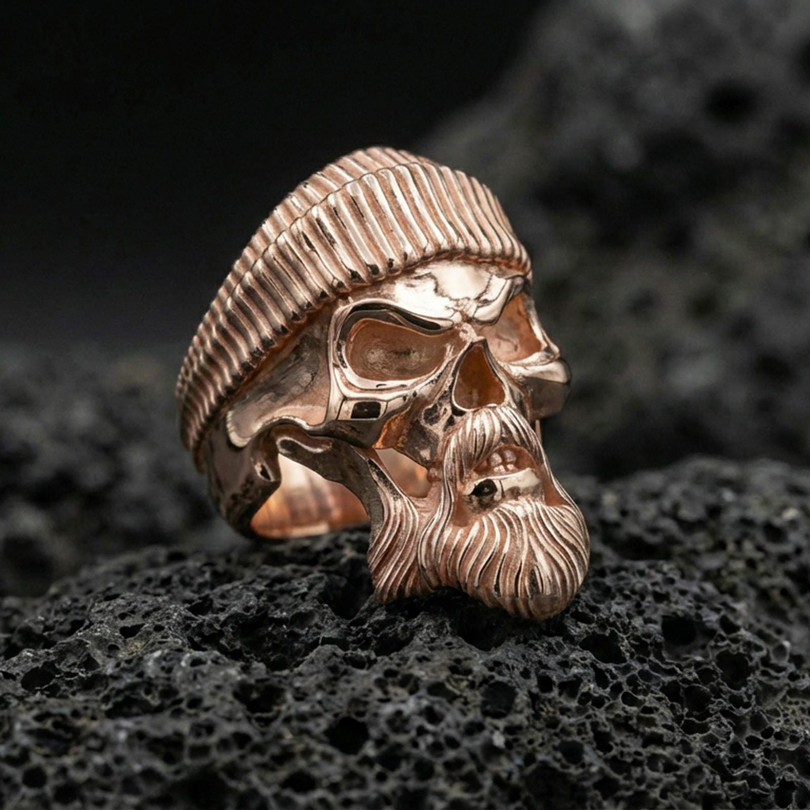 Rose gold plated bearded skull ring with ribbed beret on textured volcanic rock, luxury close-up capturing soft warm plating glow and high-relief gothic details