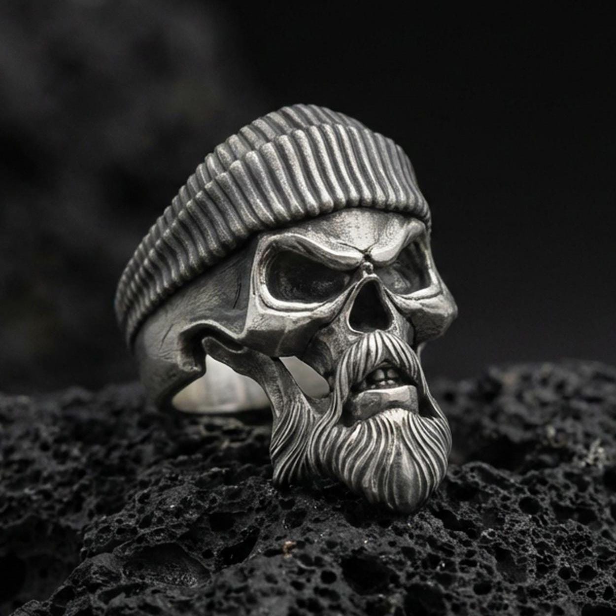 Oxidized sterling silver skull ring with ribbed beret and sculpted beard on black volcanic rock, macro close-up showing deep shadows and rugged biker detailing