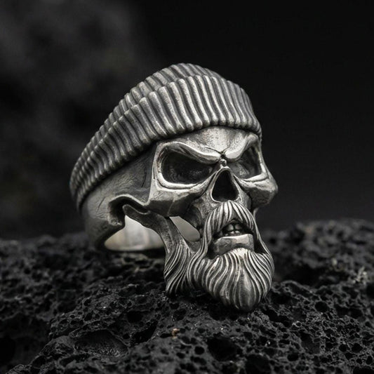 Oxidized sterling silver skull ring with ribbed beret and sculpted beard on black volcanic rock, macro close-up showing deep shadows and rugged biker detailing