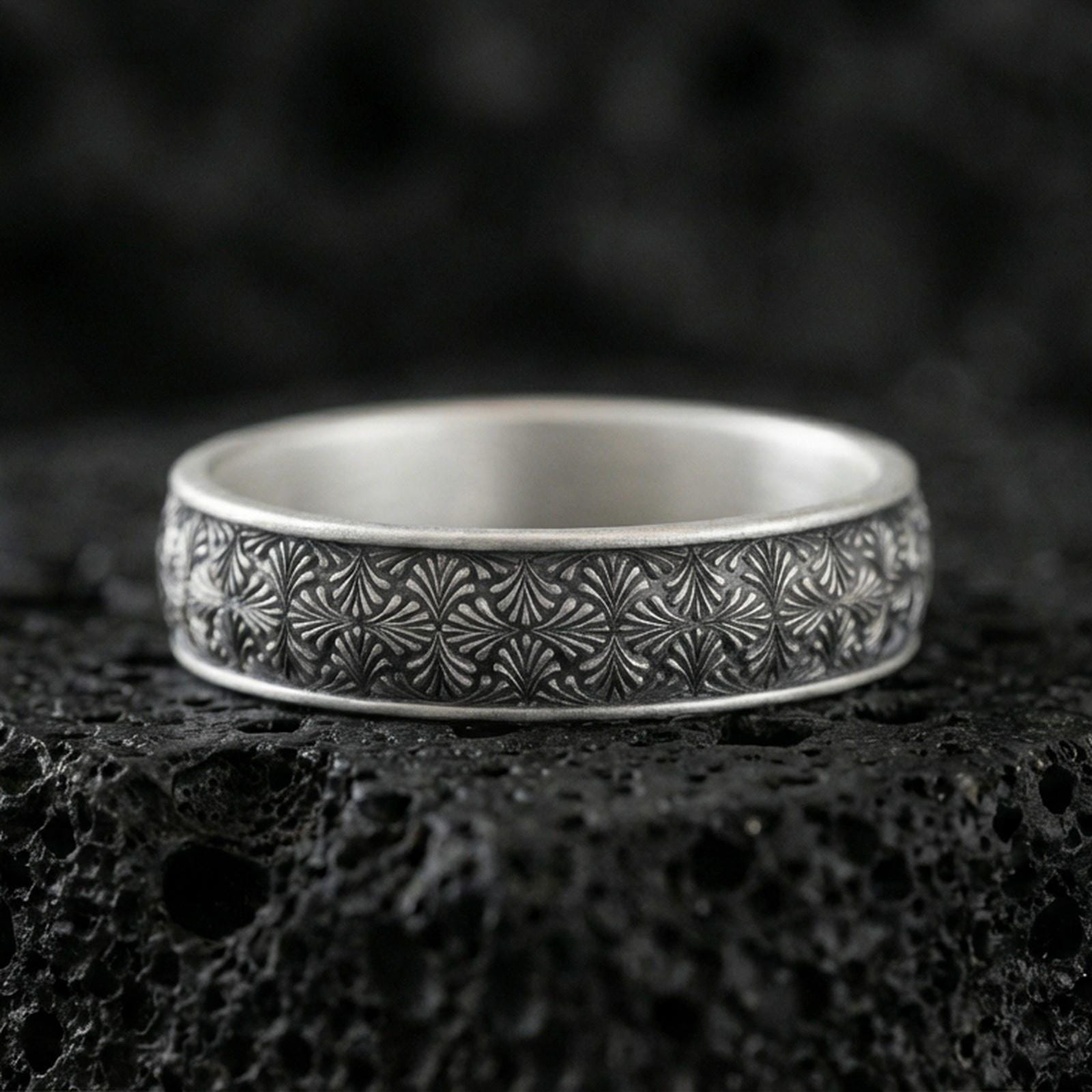 Oxidized sterling silver wedding band on dark volcanic rock, engraved starburst floral texture, premium macro product shot.