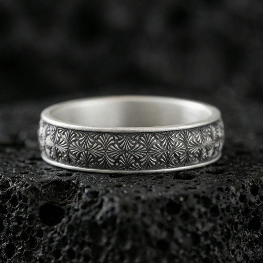 Oxidized sterling silver wedding band on dark volcanic rock, engraved starburst floral texture, premium macro product shot.