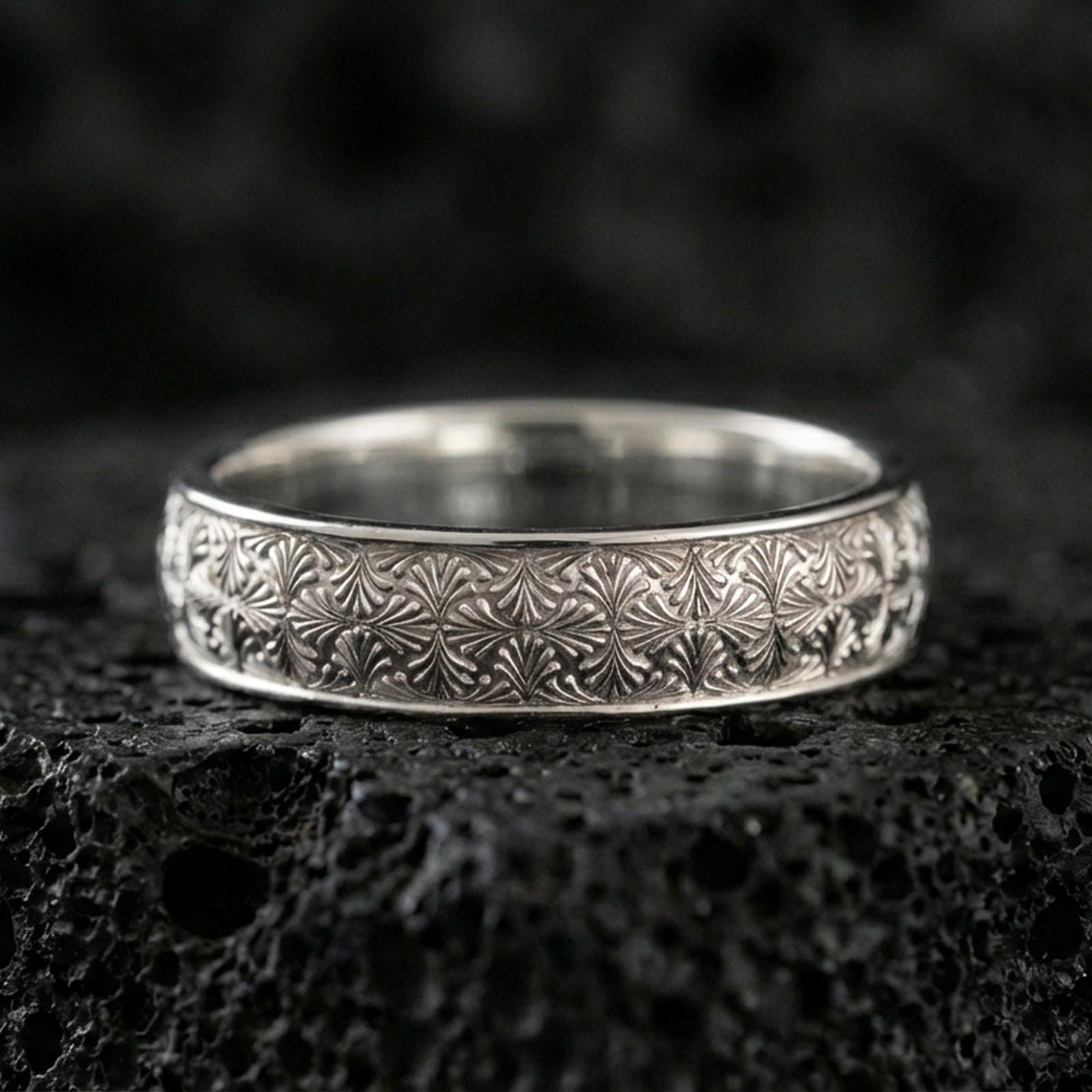 Polished silver wedding band on black volcanic stone, crisp starburst flower engraving, centered close-up studio lighting.