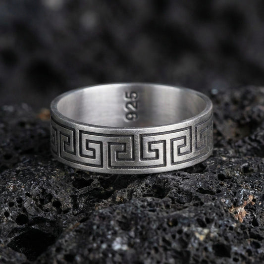 Oxidized sterling silver Greek key band ring on dark volcanic rock, bold meander pattern engraving, premium macro product shot.