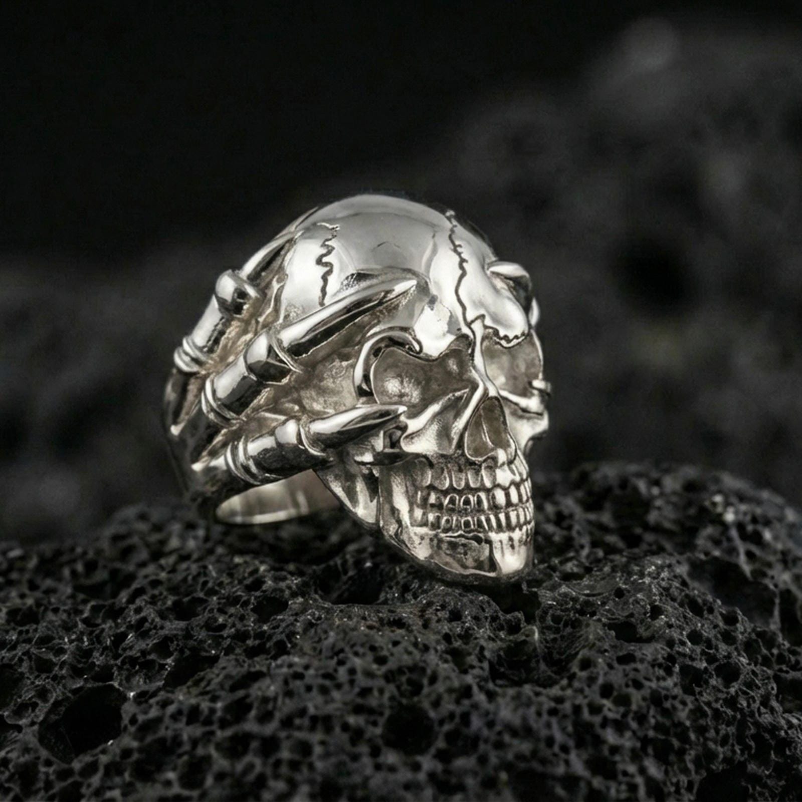 Polished silver skull ring with claw hand details on porous volcanic stone, close-up highlighting crisp teeth lines, smooth dome skull and sculpted finger bones
