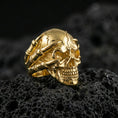 Load image into Gallery viewer, Gold plated gothic skull ring with skeletal hand grip on volcanic rock, premium macro photo emphasizing warm tone, sharp skull contours and bold biker style
