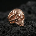 Load image into Gallery viewer, Rose gold plated skull biker ring with claw hand wrapping the skull on textured volcanic rock, luxury close-up capturing warm plating glow and high-relief details
