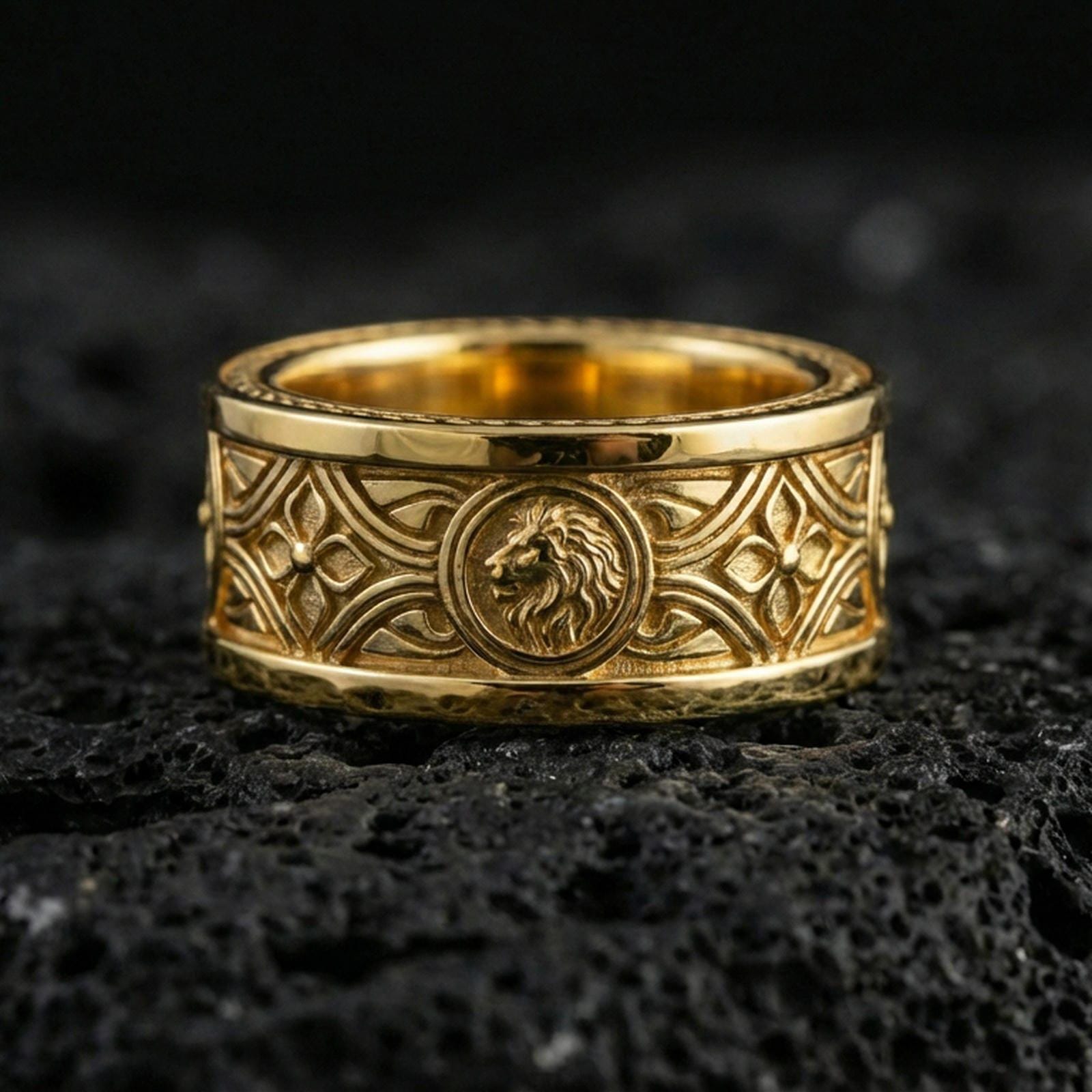 Gold plated lion medallion band ring on volcanic rock, bold central lion relief with decorative pattern borders, luxury jewelry product photography.