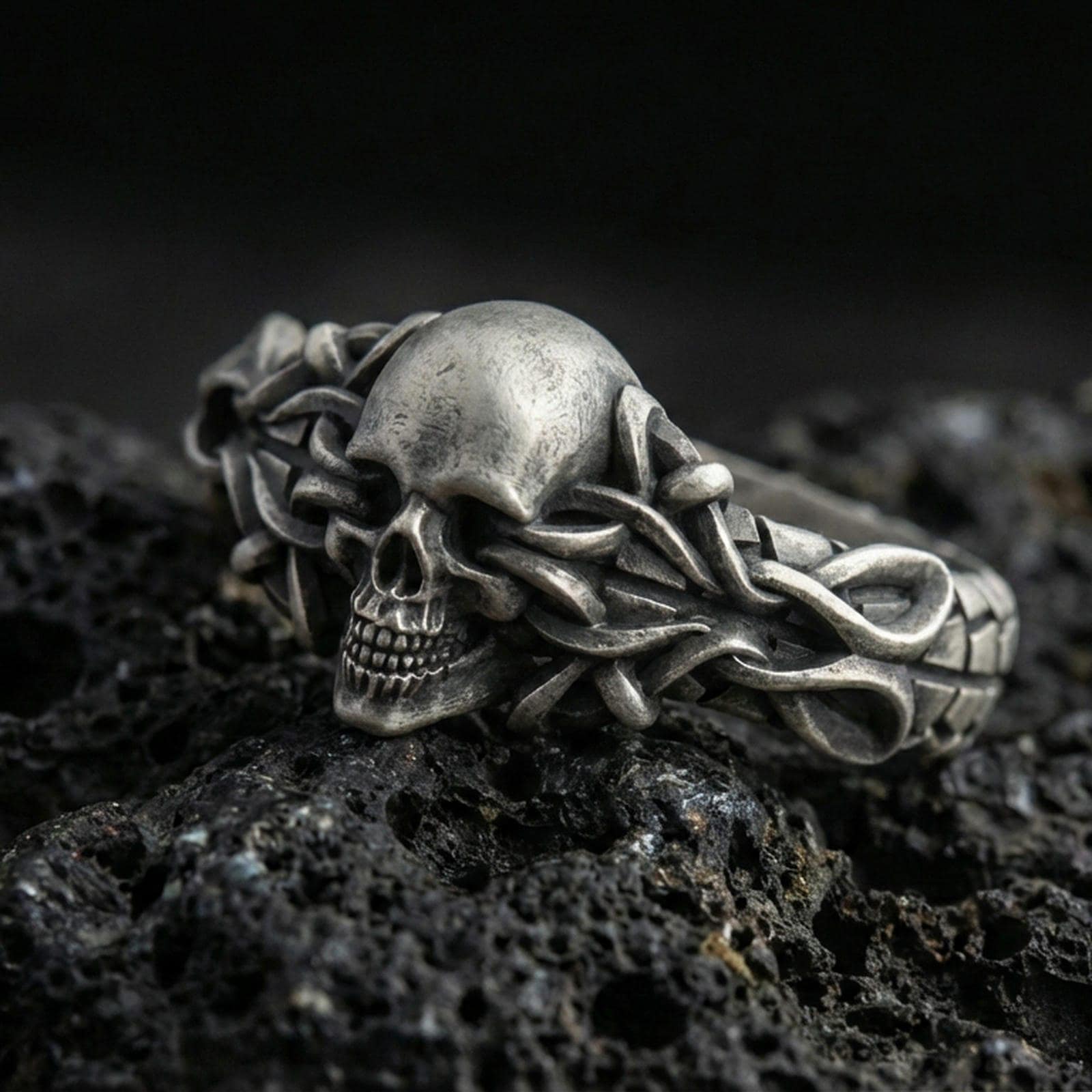 Oxidized sterling silver skull ring wrapped in thorny vine links on black volcanic rock, macro close-up emphasizing darkened recesses and gothic texture