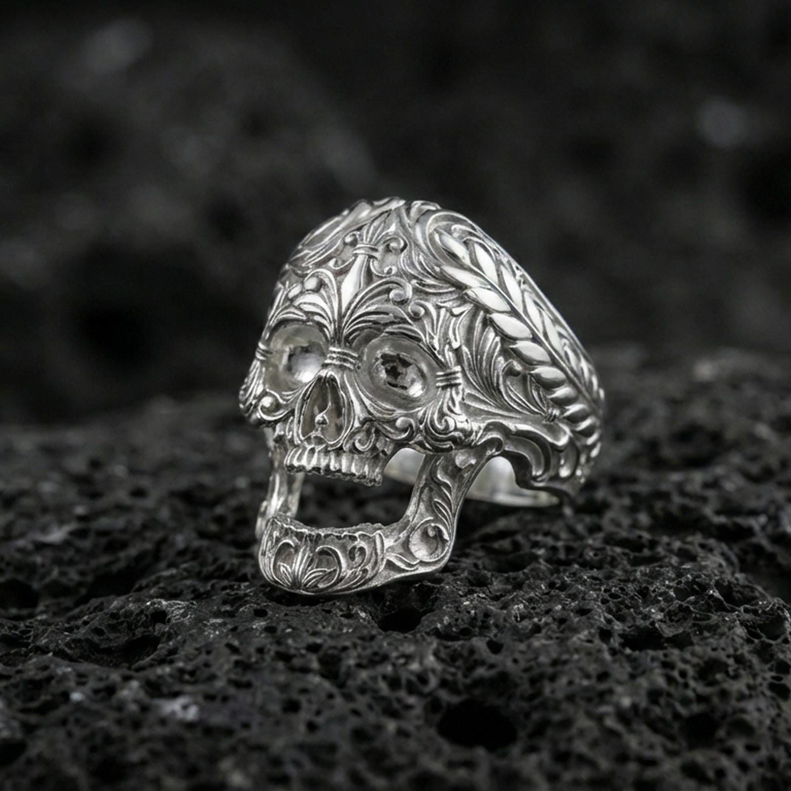 Polished silver ornate skull ring with carved scrollwork filigree and rope-edge band on porous volcanic stone, close-up highlighting crisp engraving and shine