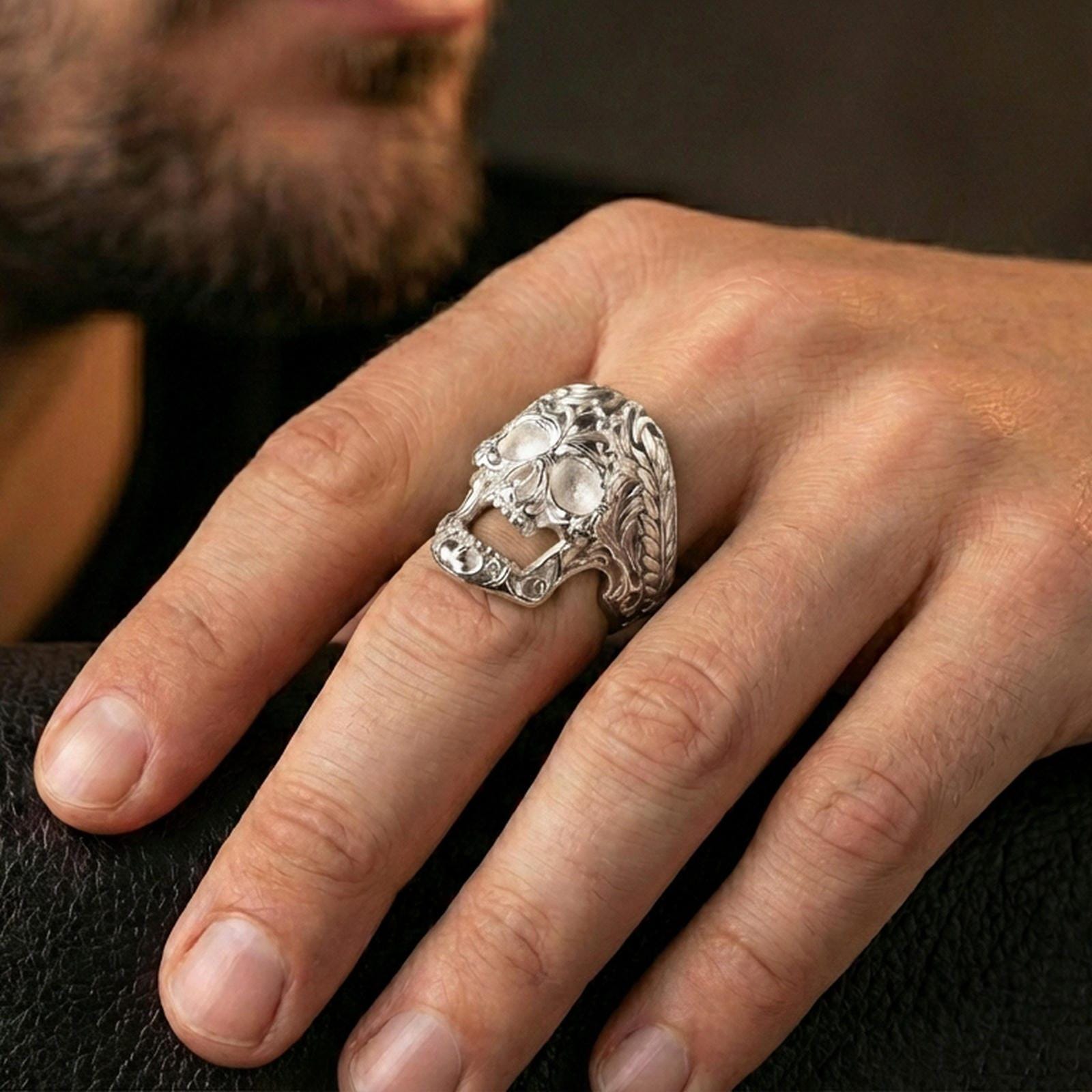 Polished silver ornate skull ring worn on the finger, wearable photo highlighting reflective finish, detailed scrollwork filigree and rope-edge band