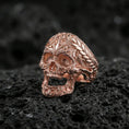 Load image into Gallery viewer, Rose gold plated ornate skull ring with floral filigree and braided edge on textured volcanic rock, close-up capturing warm plating glow and detailed skull sculpting
