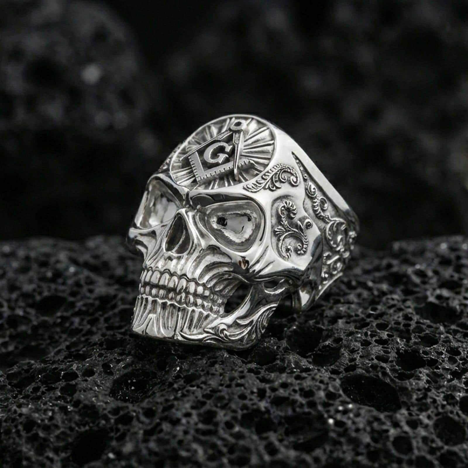 Polished silver skull ring featuring a square and compass “G” symbol on top and engraved scrollwork sides, photographed on porous volcanic stone