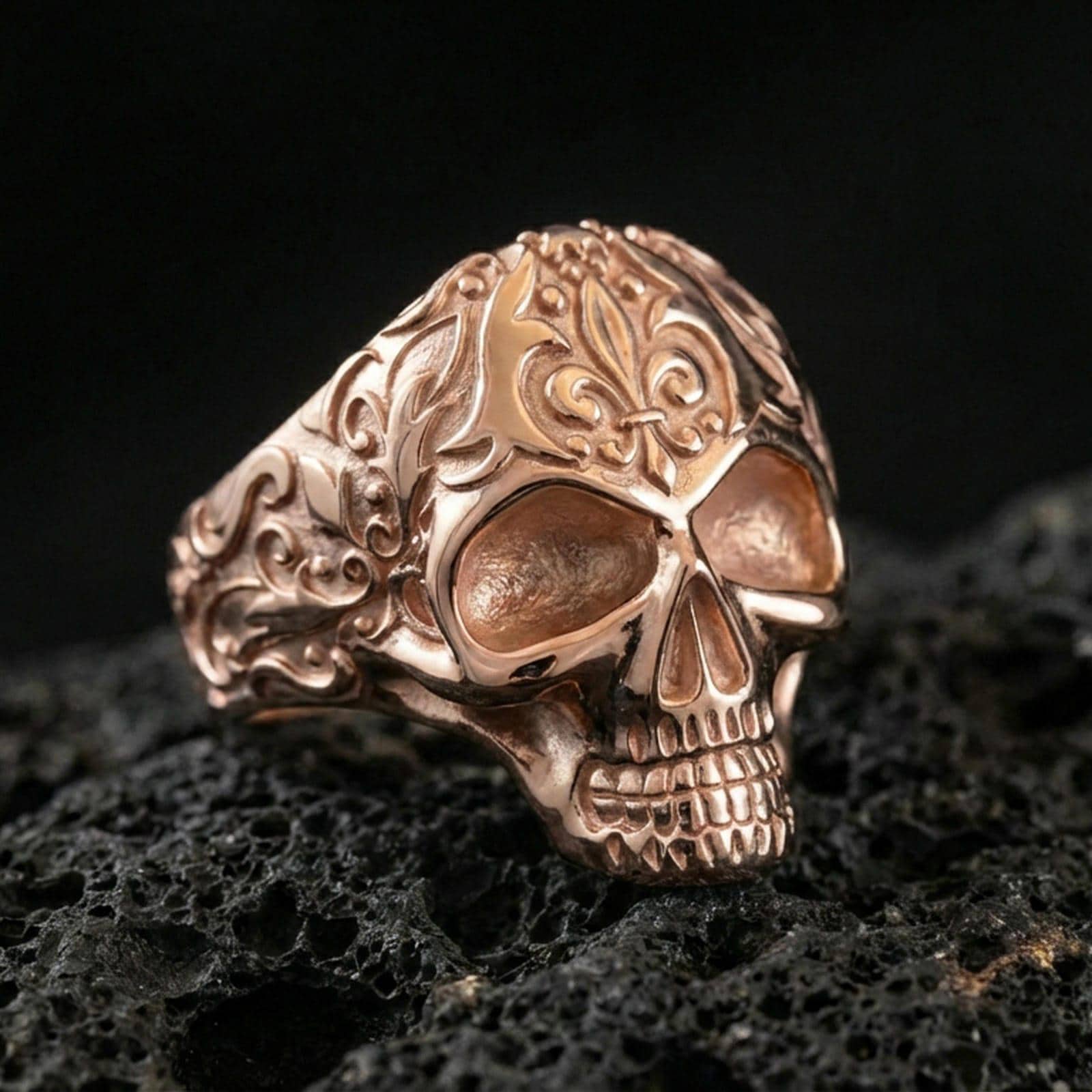 Rose gold plated skull ring with vintage floral scrollwork detailing and sculpted skull face, macro product shot on black volcanic rock