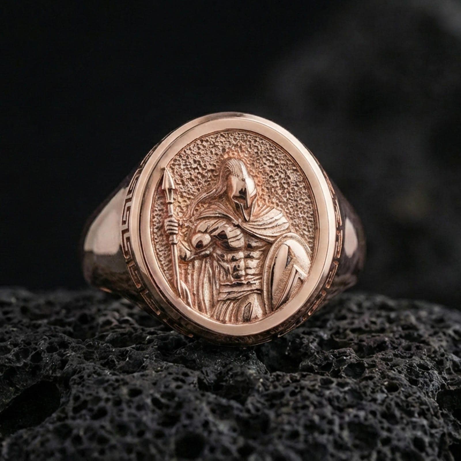 Rose gold plated Greek warrior signet ring featuring a helmeted fighter holding a spear and shield, macro product shot on black volcanic rock