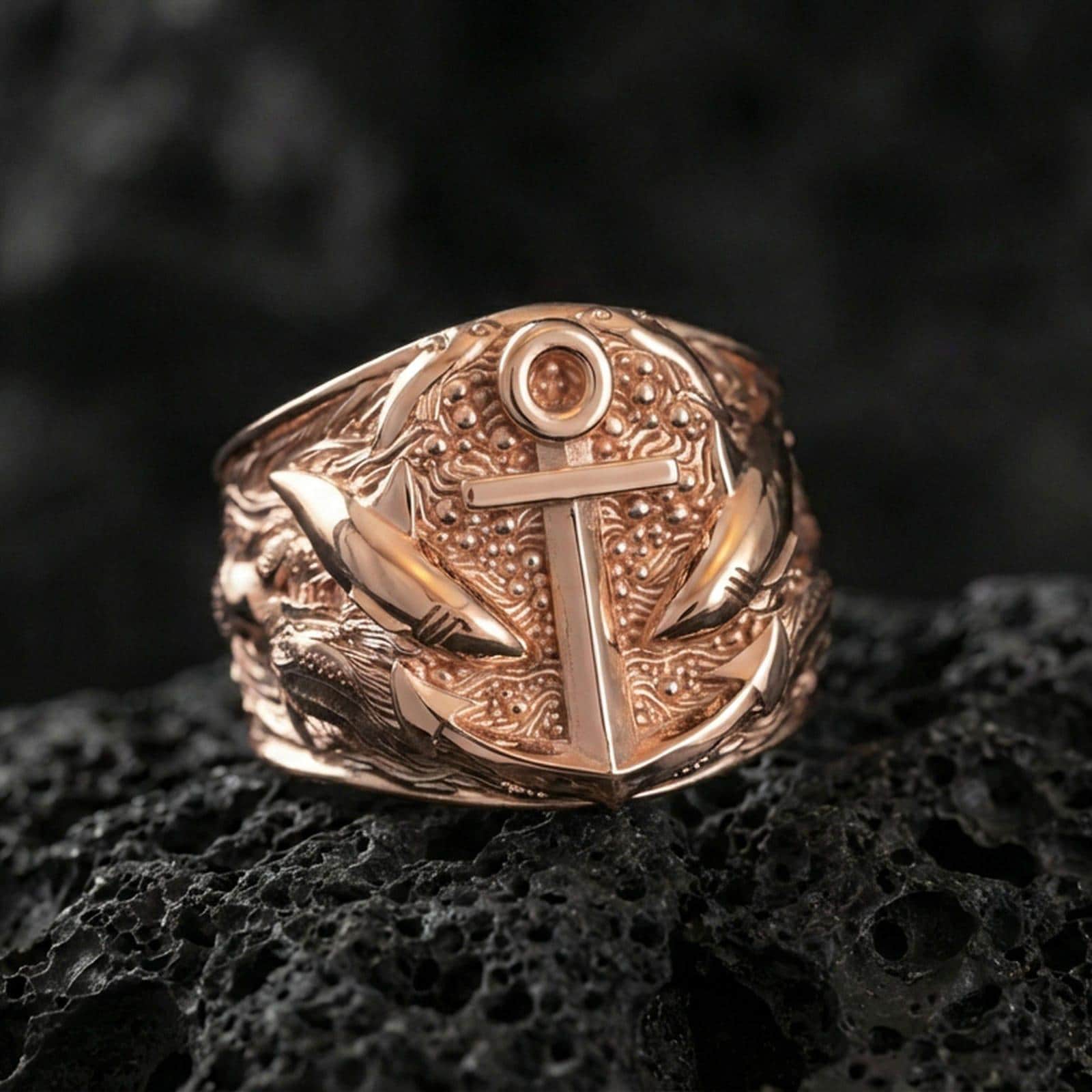 Rose gold plated nautical anchor ring with shark motif and textured ocean background, macro product shot on volcanic rock surface