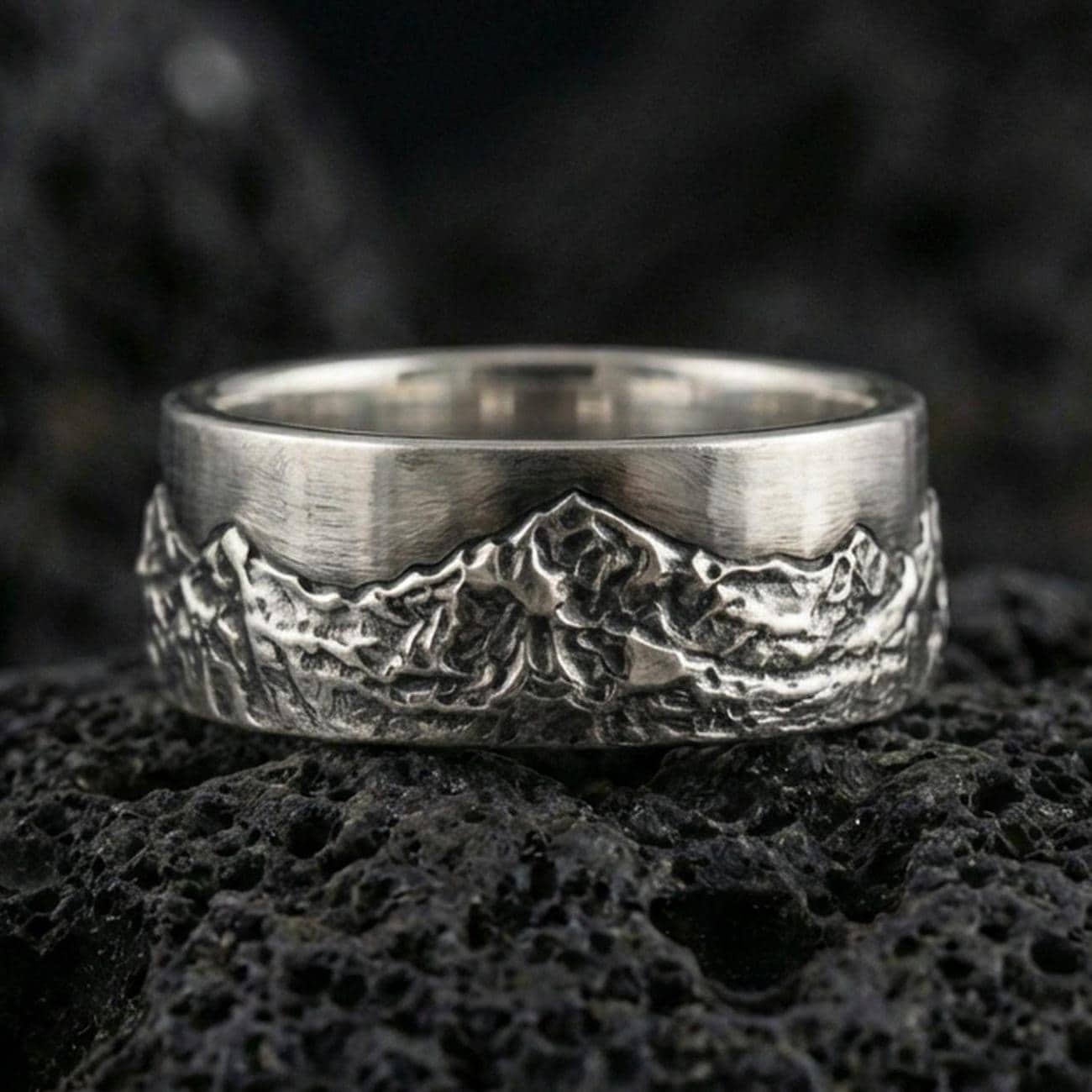 Oxidized sterling silver mountain landscape band ring on dark volcanic rock, rugged alpine relief texture, premium macro product shot.
