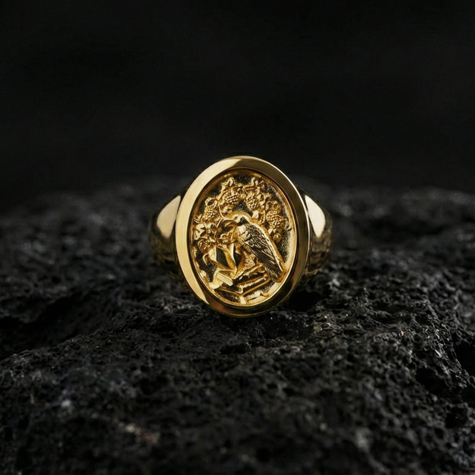 Gold plated raven crest signet ring with detailed bird engraving and decorative foliage, product-only macro image on black volcanic rock
