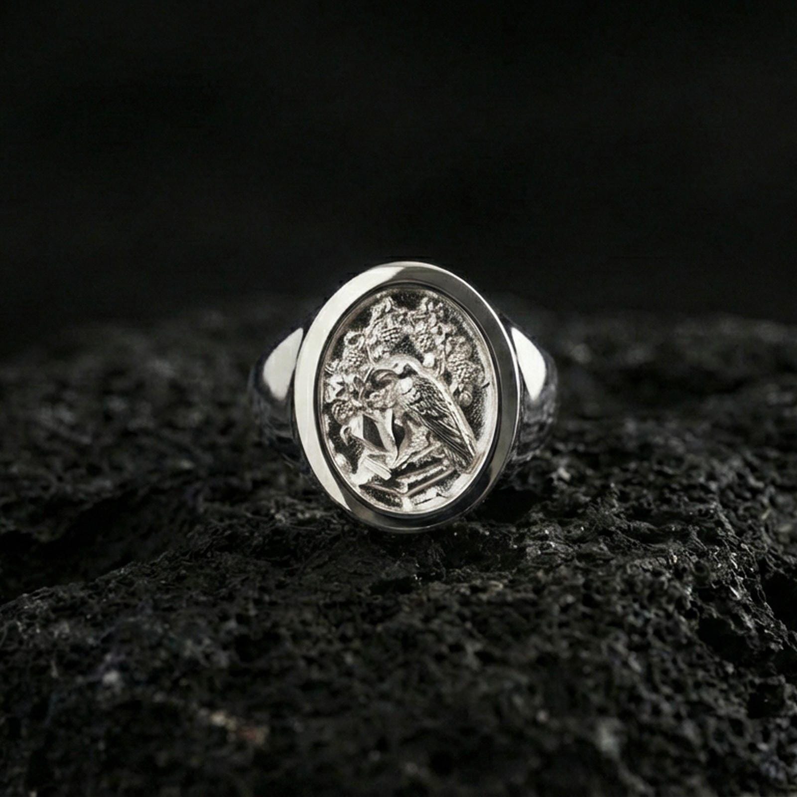 Polished silver raven signet ring featuring engraved crest details and rounded face, close-up studio shot on porous volcanic stone