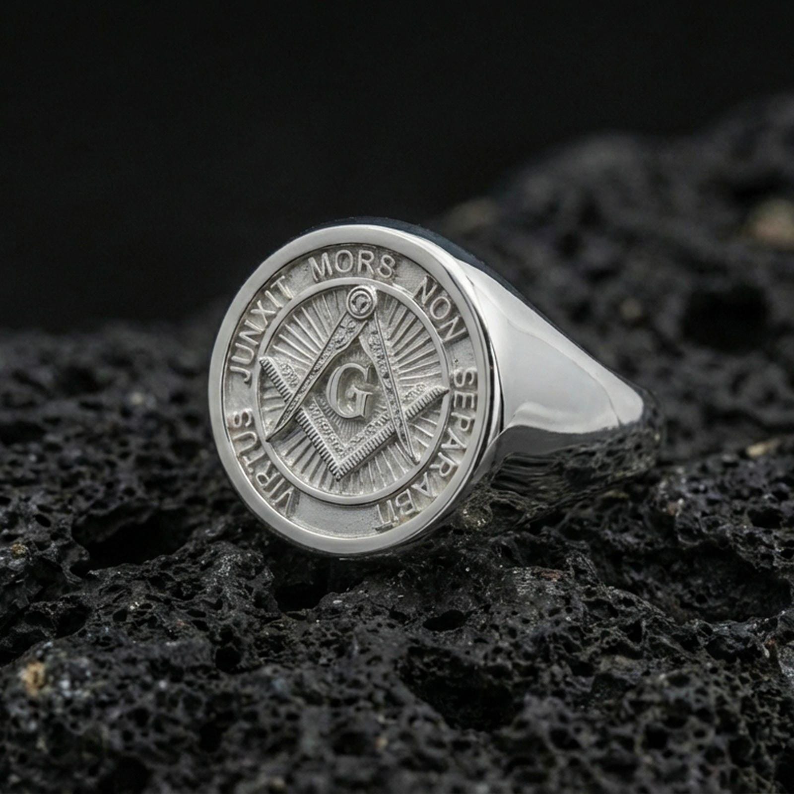 Polished silver Freemason signet ring featuring square and compass engraving with circular motto border, close-up on porous volcanic stone