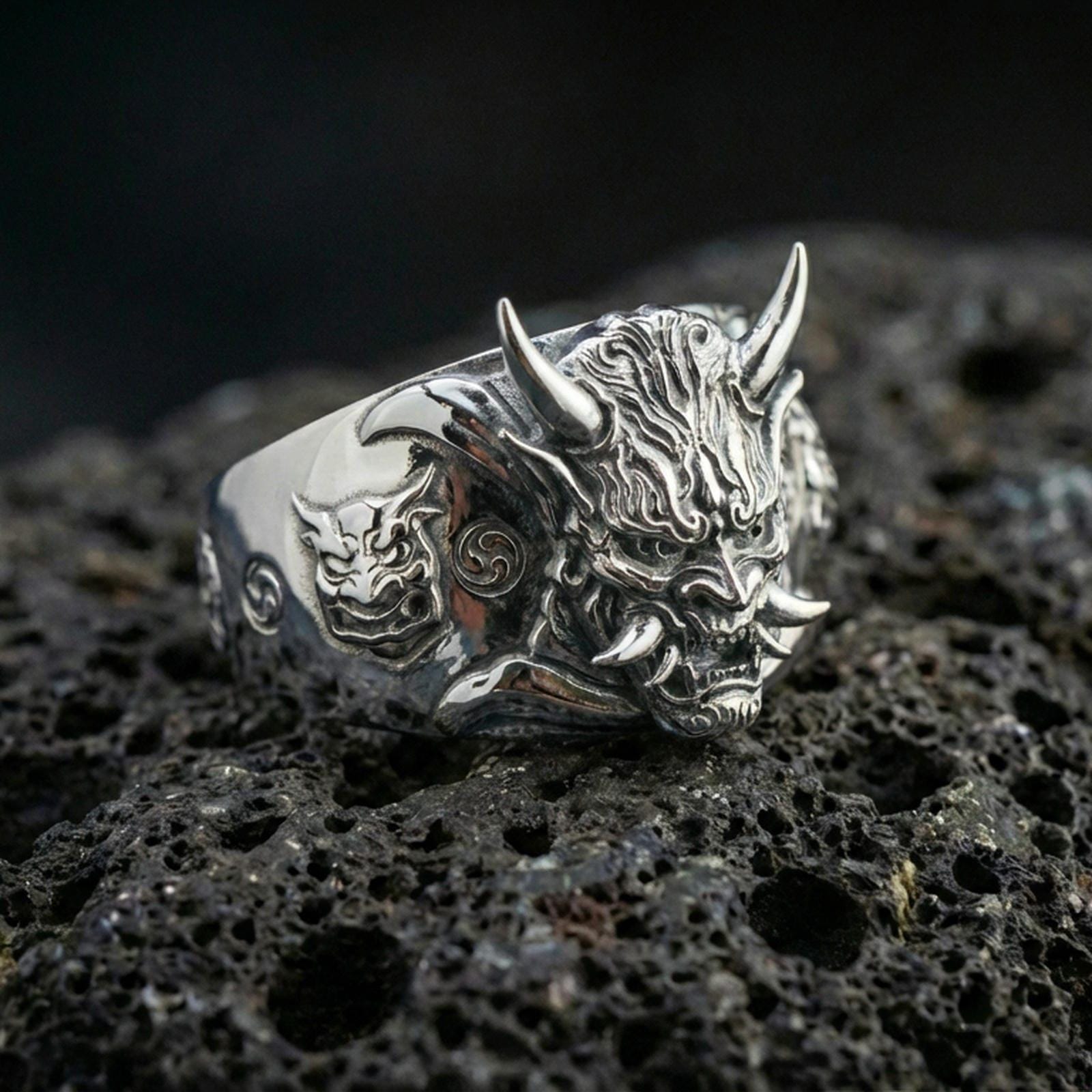 Polished silver Japanese Oni mask statement ring featuring raised horned demon head and carved swirl accents, close-up on volcanic stone