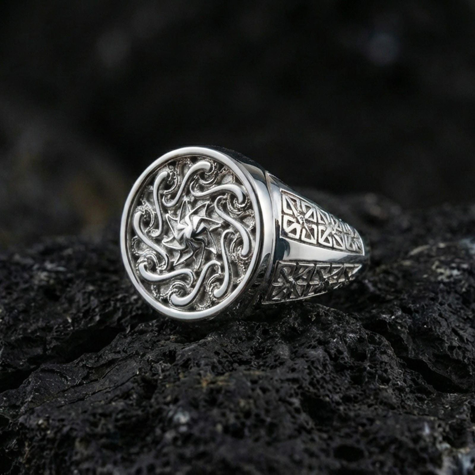 Polished silver round signet ring featuring raised spiral sun motif and engraved geometric band details, close-up on volcanic stone