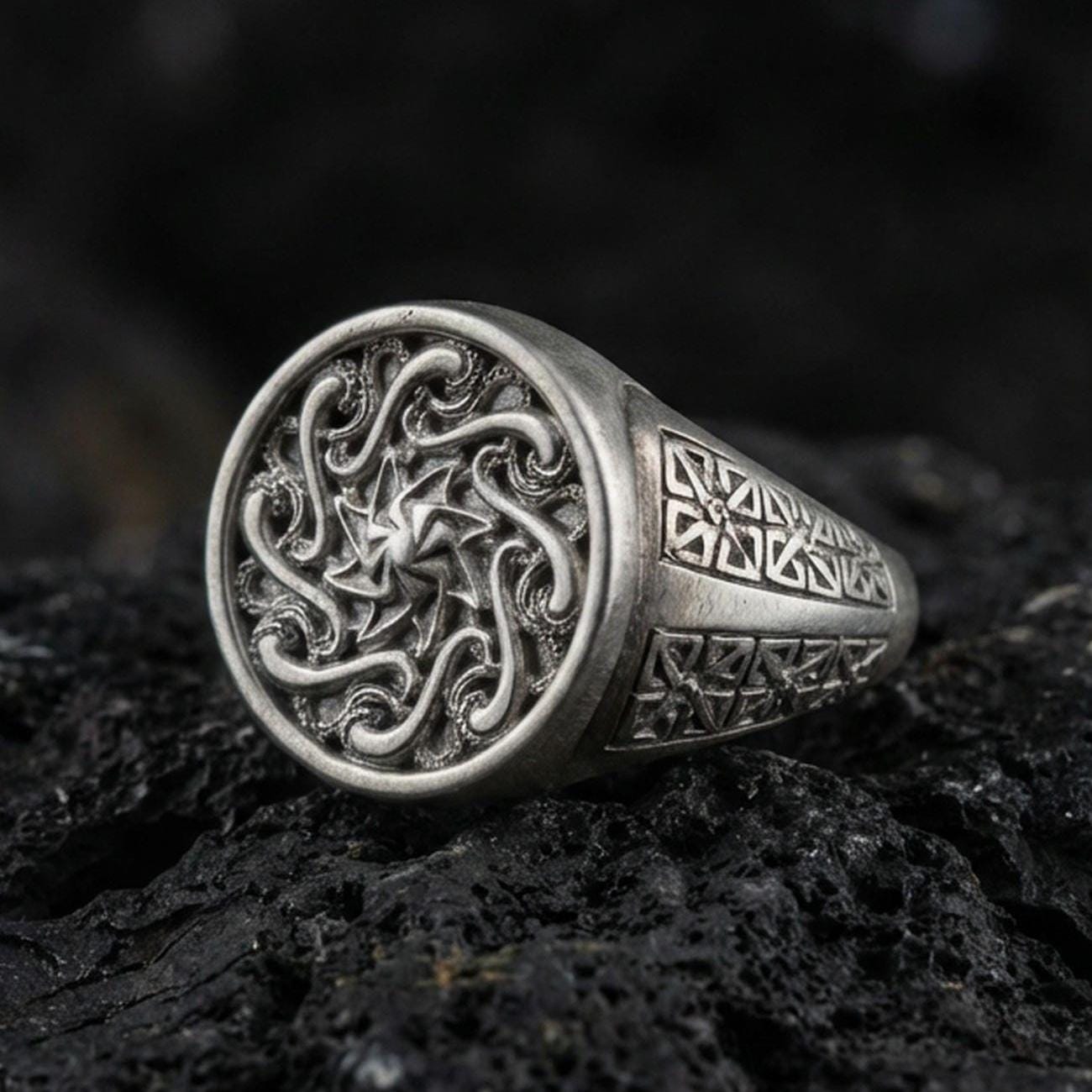 Oxidized sterling silver spiral sun signet ring with swirling Celtic-style face and geometric side panels, macro shot on black volcanic rock