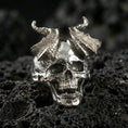 Load image into Gallery viewer, Polished silver horned skull ring featuring high-relief teeth and textured horn segments, close-up product shot on volcanic stone
