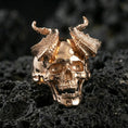 Load image into Gallery viewer, Rose gold plated horned skull ring with deep skull carving and rugged horn texture, close-up photo on volcanic rock
