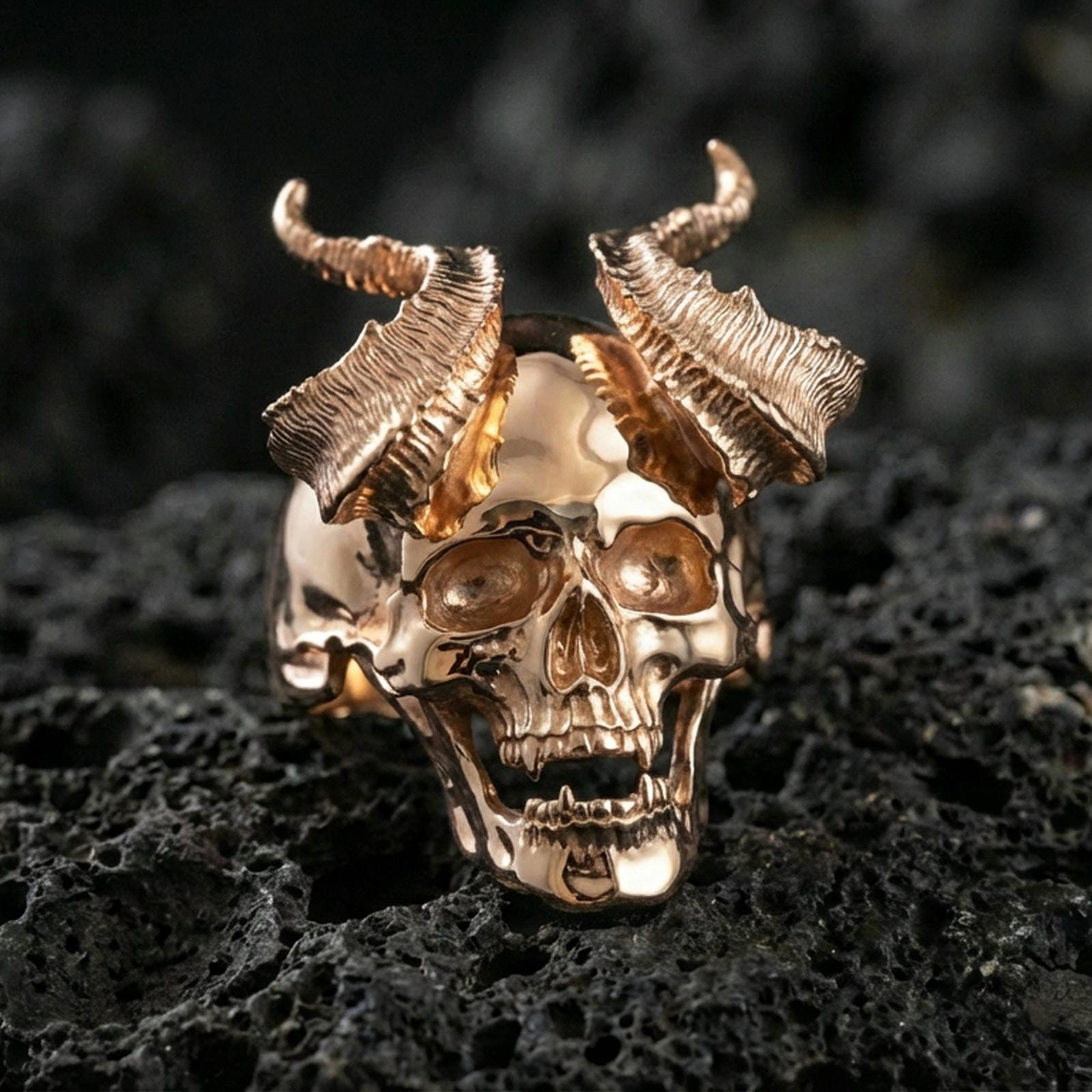 Rose gold plated horned skull ring with deep skull carving and rugged horn texture, close-up photo on volcanic rock